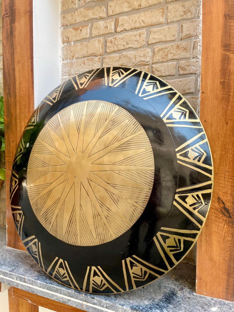 Op de afbeelding: Een groot, rond, zwart en gouden schild met een decoratief ontwerp. Het schild heeft een centraal stervormig patroon en een rand van driehoeken.
