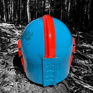 May include: A blue helmet with a red stripe down the center and red side accents. The helmet is sitting on the ground in a forest setting. The helmet appears to be made of plastic or a similar material.