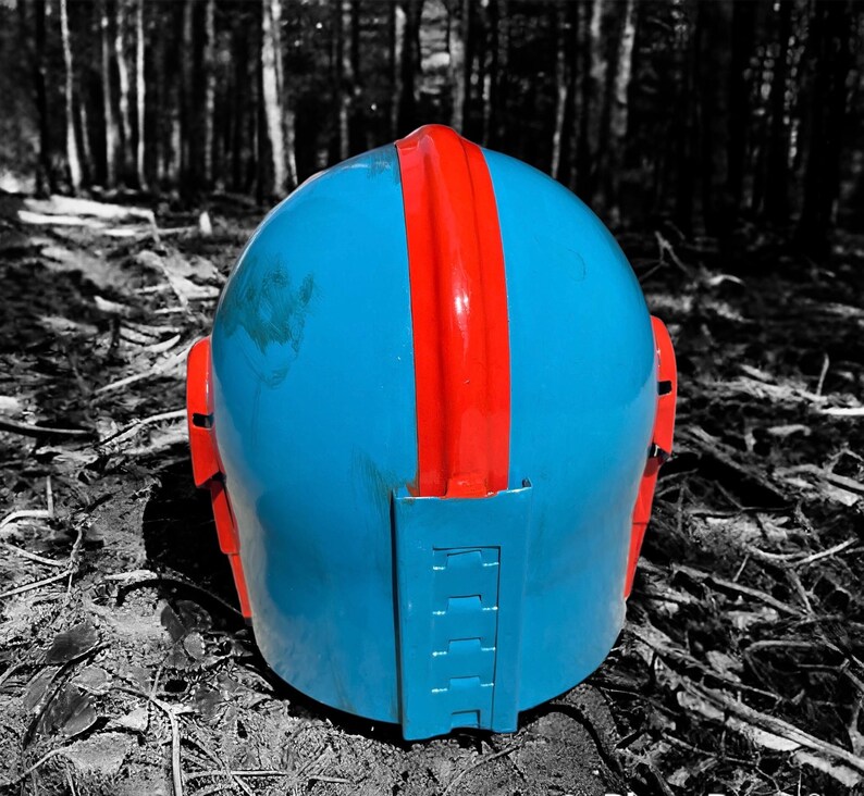 May include: A blue helmet with a red stripe down the center and red side accents. The helmet is sitting on the ground in a forest setting. The helmet appears to be made of plastic or a similar material.