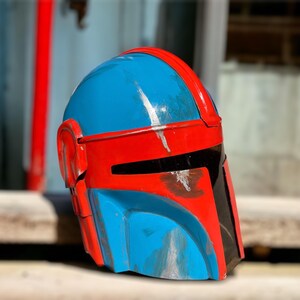 May include: A blue and red Mandalorian helmet with black visor details. The helmet has a weathered appearance with silver paint streaks. The helmet is a prop or costume piece.