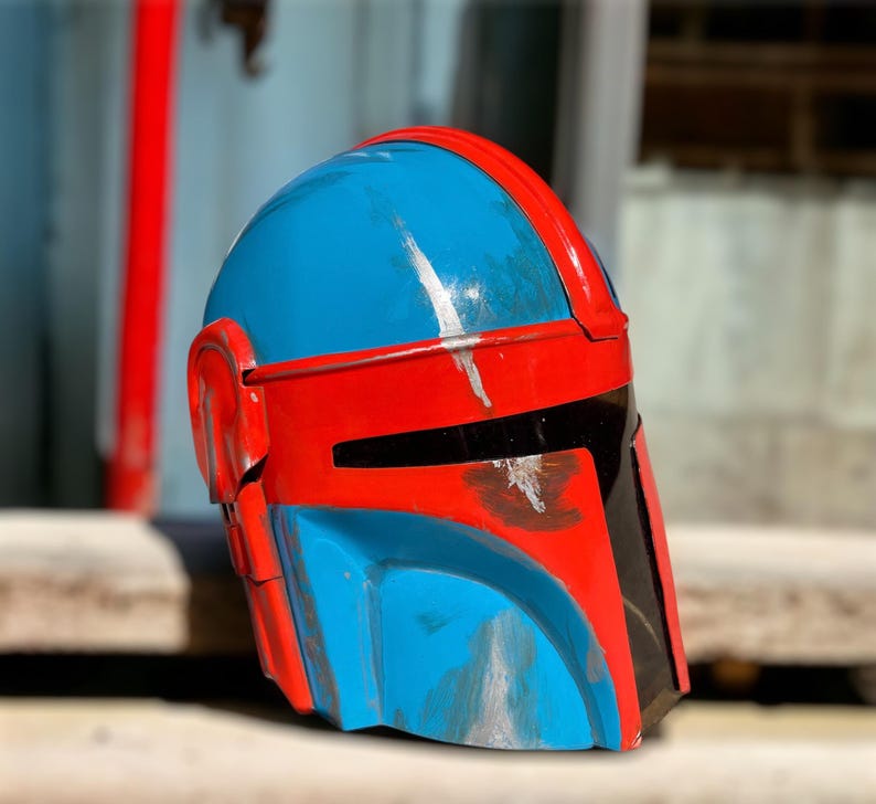 May include: A blue and red Mandalorian helmet with black visor details. The helmet has a weathered appearance with silver paint streaks. The helmet is a prop or costume piece.