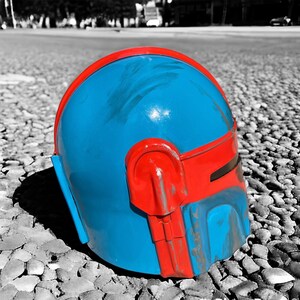 May include: A blue and red helmet, likely a costume piece, rests on a pebbled surface. The helmet features a vibrant blue dome with red accents, including a band across the forehead and side details. The color contrast is striking against the grayscale background.