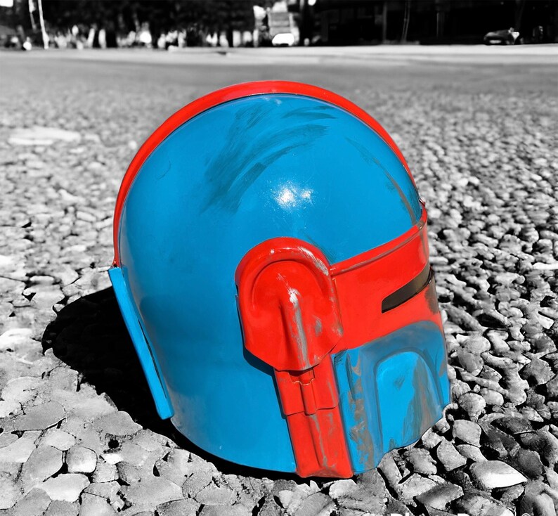 May include: A blue and red helmet, likely a costume piece, rests on a pebbled surface. The helmet features a vibrant blue dome with red accents, including a band across the forehead and side details. The color contrast is striking against the grayscale background.