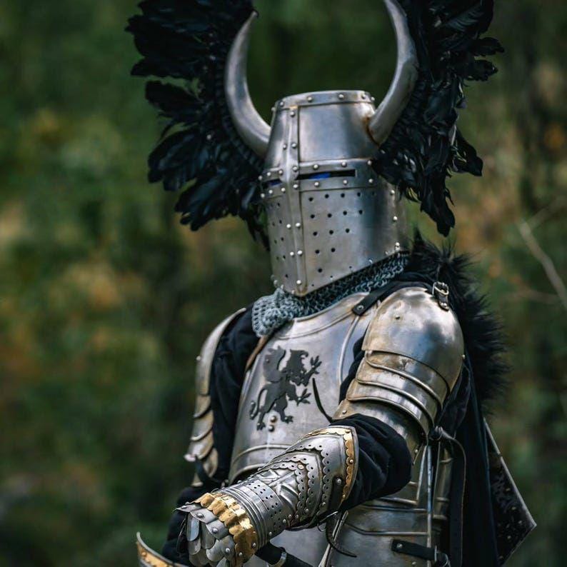 May include: A medieval knight in full armor, including a silver helmet with horns and black feather accents. The armor features a lion crest and gold detailing on the gauntlets. The knight is standing in a forest setting.