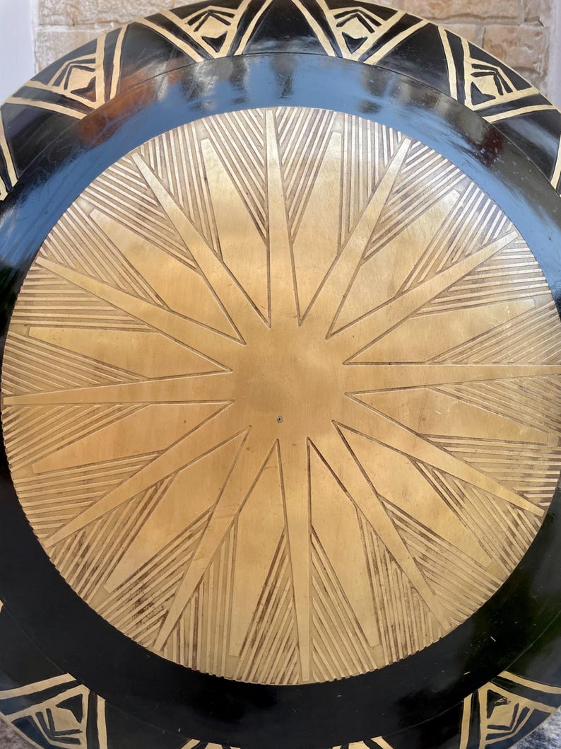 Op de afbeelding: Een rond, zwart en gouden schild met een stervormig ontwerp. Het schild heeft een zwarte buitenrand met een gouden stervorm in het midden. De stervorm is gemaakt van meerdere lijnen die vanuit het midden uitstralen.