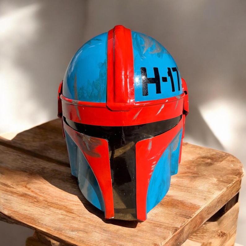 May include: A blue and red helmet with the text "H-17" on the side. The helmet has a weathered appearance with red accents and a black visor. It is sitting on a wooden surface.