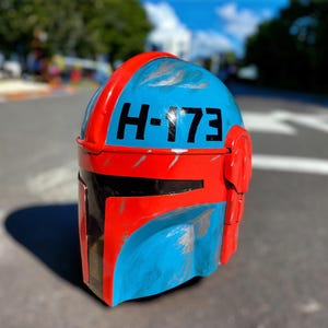 May include: A Mandalorian-style helmet in blue and red with the number "H-173" in black. The helmet has a weathered appearance with silver paint details and a black visor.