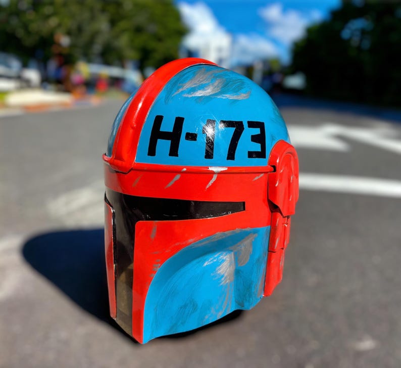 May include: A Mandalorian-style helmet in blue and red with the number "H-173" in black. The helmet has a weathered appearance with silver paint details and a black visor.