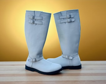 May include: A pair of light gray riding boots with black soles. Each boot features two sets of buckles at the top and one at the ankle. The boots are made of a smooth material and stand on a wooden surface.