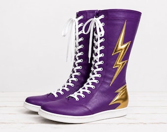 May include: A pair of mid-calf purple boots featuring white lace-up closures, a flat white sole, and a metallic gold lightning bolt appliqué on the outer side. The boots have a smooth finish and a rounded toe design with a gold accent at the heel.