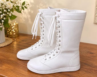 Pode incluir: Um par de botas brancas de cano médio com acabamento liso, biqueiras arredondadas e atacadores frontais completos com cordões brancos e ilhós prateados. As botas têm uma sola branca plana e um design de cano alto estruturado.