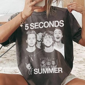May include: Dark gray t-shirt featuring the text "5 SECONDS OF SUMMER" above a graphic of five young men. The shirt is a relaxed fit and appears to be made of a soft material. The shirt is worn with gray shorts and white socks with black stripes.