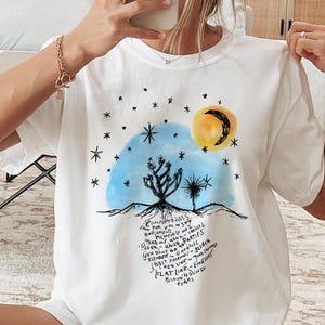 May include: White t-shirt with a desert landscape design. The graphic features a blue sky with stars, a yellow moon, and a Joshua tree. The text on the shirt reads "COMPLETE MESS" and other phrases.