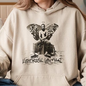 May include: Beige hoodie featuring a black and white graphic of a person sitting with a crown, surrounded by a crowd. The text "Kendrick Lamar" is printed below the graphic in a handwritten style. The hoodie has a front pocket and a drawstring hood.