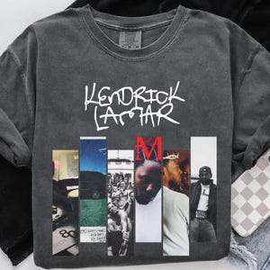 May include: Dark gray sweatshirt featuring the text "Kendrick Lamar" in white script above a series of vertical images. The letter "M" is in red. The sweatshirt has a vintage, washed appearance.