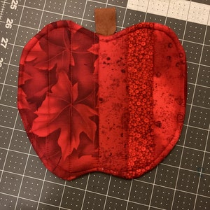 May include: Red quilted apple-shaped hot pad with a brown leather loop. The hot pad is made of fabric with a maple leaf pattern on one side and a red and white polka dot pattern on the other side.