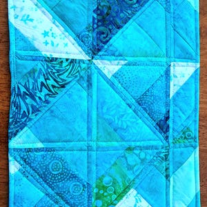 May include: A rectangular quilted fabric mat featuring a geometric patchwork design in various shades of blue, turquoise, and teal. The surface displays a mix of solid and patterned fabric triangles and squares with visible straight-line stitching.
