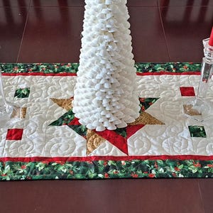 May include: A rectangular Christmas table runner with a quilted design. The runner features a white background with red, green, and gold star patterns. A green border with red berries surrounds the design. Two clear glass candle holders with red candles and a white tree are on the runner.