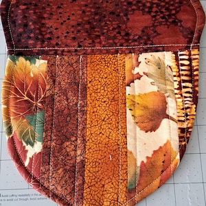 May include: A quilted fabric potholder shaped like an acorn, featuring a dark brown textured top and a lower section composed of vertical fabric strips in shades of orange, yellow, and brown with printed autumn leaf and crackle patterns.
