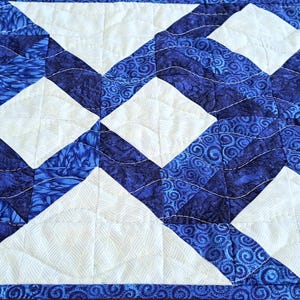 May include: A quilted table runner featuring a repeating pattern of white diamond shapes set against a backdrop of various blue patterned fabrics. The runner has a blue border and is likely handmade.
