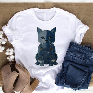 May include: White t-shirt featuring a cat illustration wearing denim overalls. The cat is split down the middle with one side gray and the other blue. A pair of blue jeans and tan boots are next to the shirt.