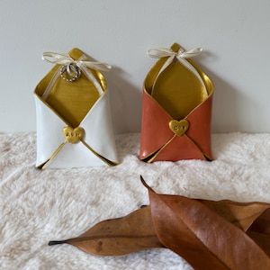 May include: Two envelope-shaped holders, one white and one terracotta, each featuring a gold interior, a gold heart-shaped clasp, and a decorative ribbon bow at the top. The white holder displays two silver rings hanging from the ribbon bow.