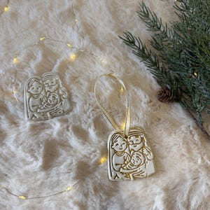 Peut inclure: Deux ornements plats de couleur blanc cassé, moulés avec un motif en relief représentant une famille de trois personnes. L'un des ornements possède une fine boucle de ruban crème fixée au sommet, tandis que l'autre est posé à plat.