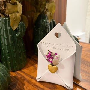 Pode incluir: Um envelope de papel branco dobrado com um recorte em forma de coração e texto em relevo. Dentro, um pequeno buquê de flores secas rosa e roxas e um pingente de coração dourado. Sobre uma superfície de madeira.