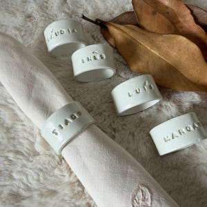 Pode incluir: Uma toalha de linho creme é circundada por quatro argolas de cerâmica branca com nomes e uma quinta argola na toalha. As argolas têm nomes em relevo. Folhas castanhas repousam sobre a superfície texturizada.