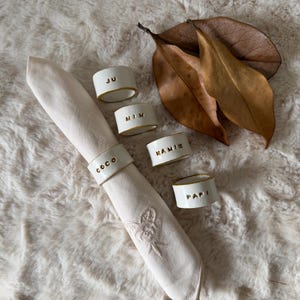 Pode incluir: Uma toalha de mesa creme com um desenho floral é segurada por argolas de guardanapo de cerâmica branca. Cada argola tem uma borda dourada e um nome em letras douradas: COCO, JU, MIM, MAMIE e PAPI. Duas folhas marrons secas grandes estão no fundo.