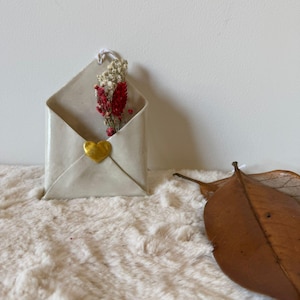 Könnte beinhalten: Eine cremefarbene Keramik-Briefumschlag mit einem goldenen Herzsiegel, das getrocknete rote und weiße Blumen enthält. Der Umschlag liegt auf einer flauschigen, weißen Oberfläche. Ein braunes Blatt liegt in der Nähe.