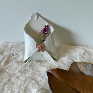 Op de afbeelding: Een witte keramische envelopvormige wandvaas met een roze strik en gedroogde bloemen. De vaas heeft een textuuroppervlak en een lus om op te hangen. De achtergrond is een witte muur en een zachte witte vloerkleed.