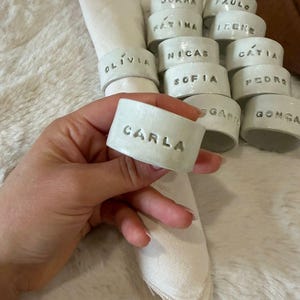 Può includere: Anelli portatovaglioli in ceramica bianca, ognuno con un nome in lettere in rilievo. Gli anelli sono presentati con un tovagliolo bianco piegato. Un anello, tenuto in mano, porta il nome "CARLA". Il design suggerisce un tema da tavola o da pranzo.