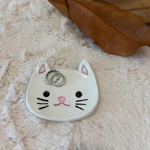 Può includere: Un piattino in ceramica bianca a forma di gatto con occhi, baffi neri e naso rosa dipinti. Le orecchie presentano accenti rosa e due anelli in metallo color argento poggiano sulla superficie del piattino, caratterizzato da una finitura lucida.