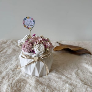 Op de afbeelding: Een witte geometrische vaas gevuld met roze, witte en paarse bloemen. Een klein houten hart en een touwstrik sieren de vaas. Een bloemrijk versierde kaart op een draadsteel is in de opstelling geplaatst. De tekst op de kaart luidt "Feliz dia da Madrinha".