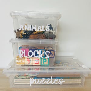 May include: Three clear plastic storage bins with labels. The top bin is labeled "ANIMALS" and contains plastic animal figurines. The middle bin is labeled "BLOCKS" and contains wooden blocks. The bottom bin is labeled "puzzles" and contains a stack of puzzles.