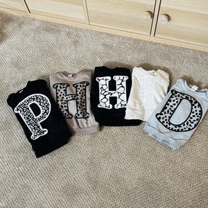 May include: Five folded sweatshirts with large letter designs. The letters are in various styles, including a black sweatshirt with a white polka dot "P", a tan sweatshirt with a brown leopard print "H", and a black sweatshirt with a white cracked design "H".