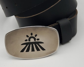 Puede incluir: Un cinturón de cuero negro con una hebilla plateada que presenta un sol estilizado que sale sobre un campo de trigo.