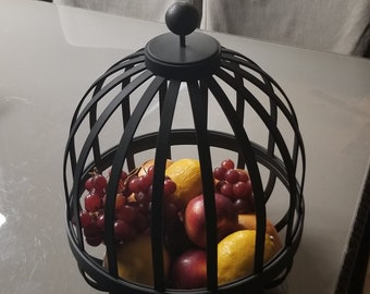 May include: Black metal fruit bowl with a dome-shaped lid. The bowl is filled with red grapes, yellow lemons, and red apples.