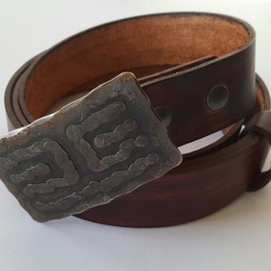 May include: A brown leather belt with a unique, textured silver buckle. The buckle features an abstract design. The belt is rolled up, showcasing the rich color and quality of the leather. A stylish accessory.