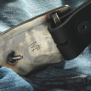 May include: A close-up of a silver metal belt buckle with a leather strap. The buckle has a small metal pin and the inscription "B.P." on the front.