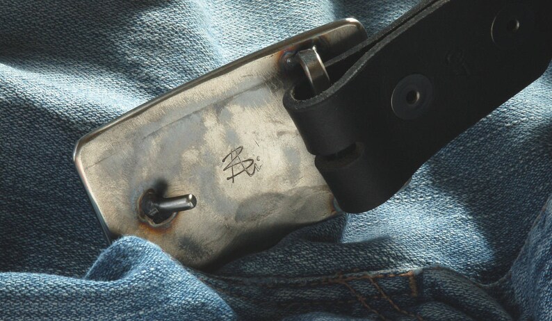 May include: A close-up of a silver metal belt buckle with a leather strap. The buckle has a small metal pin and the inscription "B.P." on the front.