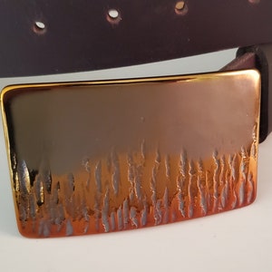 May include: A dark brown leather belt featuring a rectangular metal buckle with a polished gold-tone finish. The lower portion of the buckle displays a textured, vertical, flame-like pattern that transitions from a bright copper to a dark bronze.