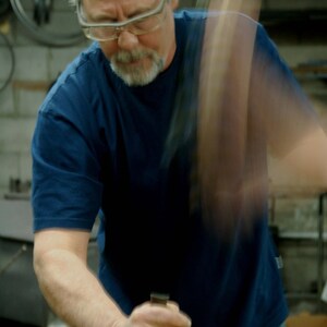 May include: A man wearing glasses and a blue t-shirt is working with a metal object. He is using a tool to shape the metal.