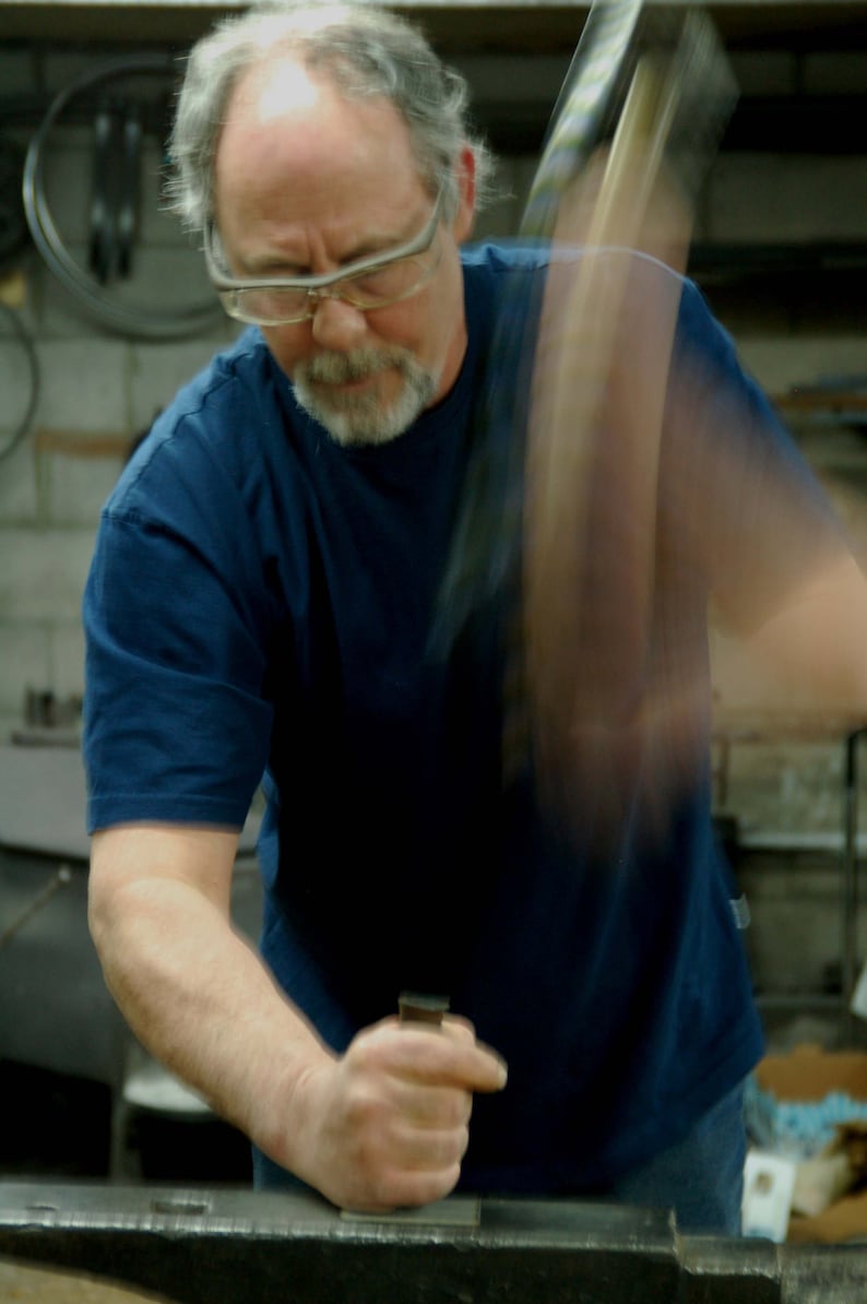 May include: A man wearing glasses and a blue t-shirt is working with a metal object. He is using a tool to shape the metal.