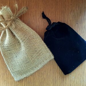 May include: Two small bags on a wooden surface. One is made of tan burlap and tied with twine. The other is a black velvet pouch with a drawstring closure. Both bags are empty and ready for use.