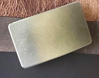 May include: A rectangular, silver-colored metal belt buckle with rounded corners. The buckle has a textured surface and a polished edge. The background features a brown, textured leather surface.
