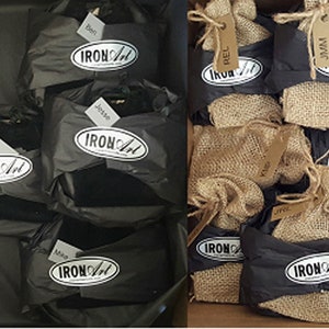 May include: A close-up of a box filled with small black gift bags and burlap sacks. Each bag has a white oval label with the text "IRON ART" printed on it. The bags are labeled with names, such as "Bill", "Jesse", "David", "Rel", and "Jam".