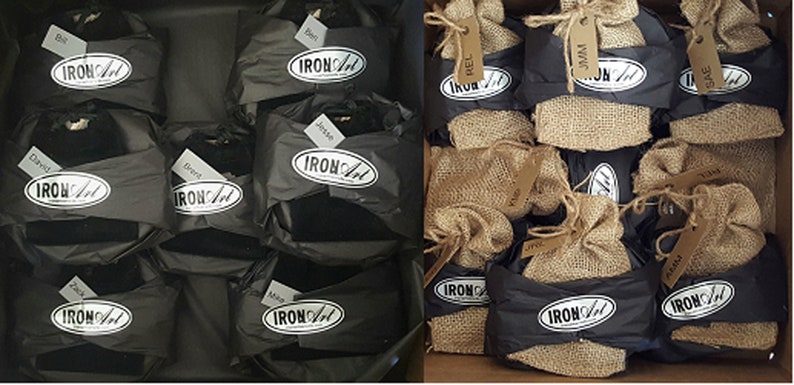 May include: A close-up of a box filled with small black gift bags and burlap sacks. Each bag has a white oval label with the text "IRON ART" printed on it. The bags are labeled with names, such as "Bill", "Jesse", "David", "Rel", and "Jam".