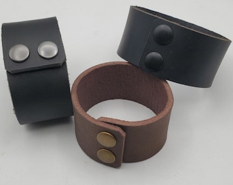 May include: Three leather wrist cuffs in black and brown. The black cuffs have silver or black snap closures. The brown cuff has a gold snap closure. The cuffs are made of thick leather.